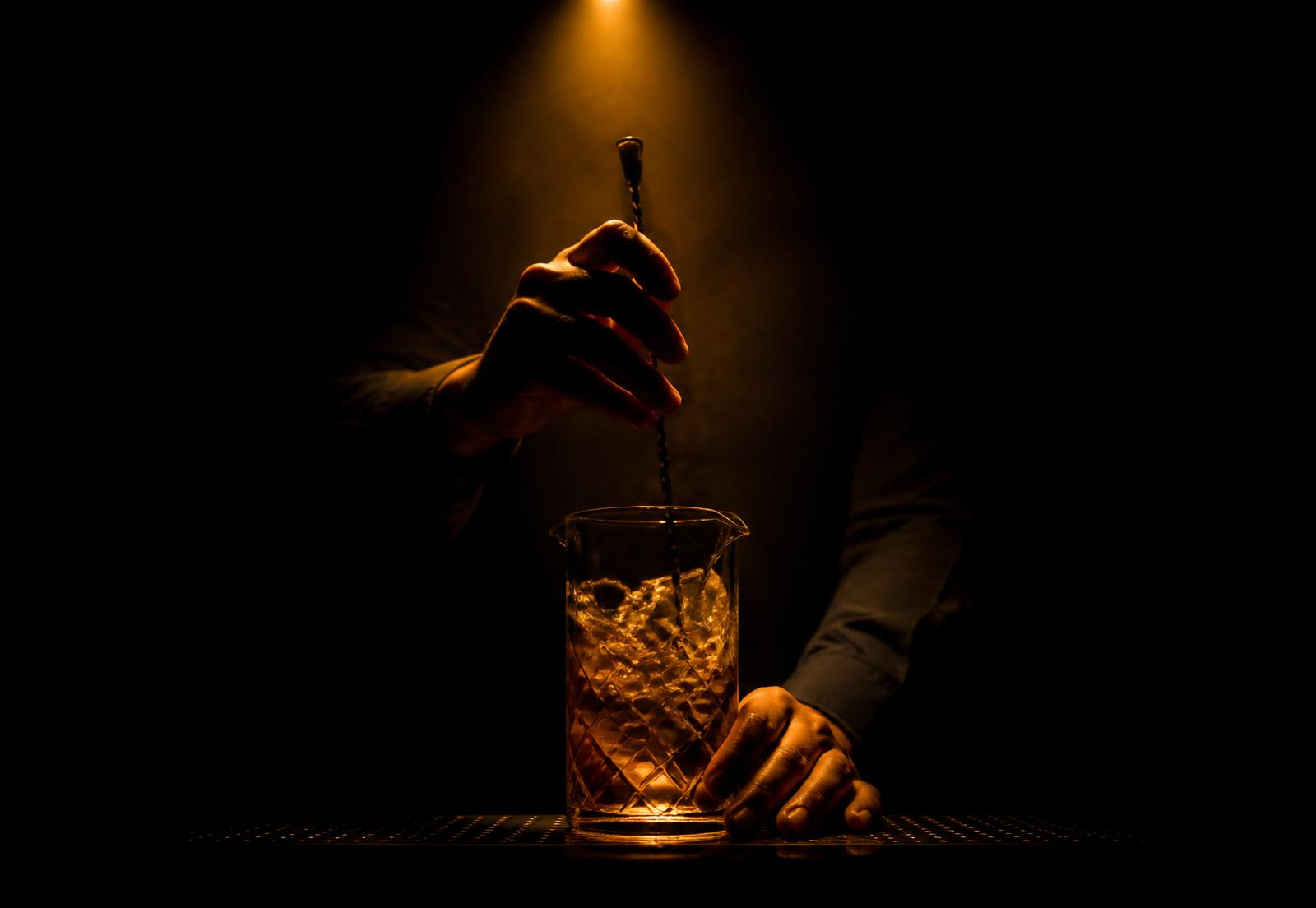 Silhouetted hands stirring a cocktail in a mixing glass under warm light